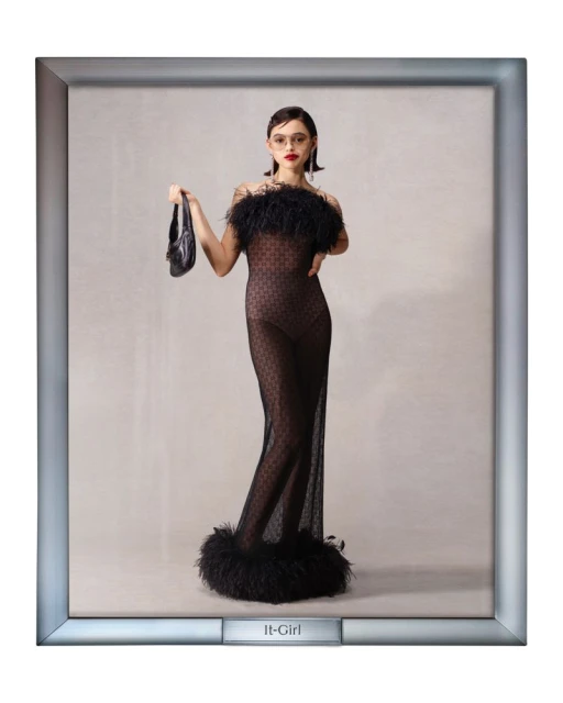 Elegant woman poses in a black sheer dress with feather trim, holding a black purse, set against a neutral background inside a picture frame.