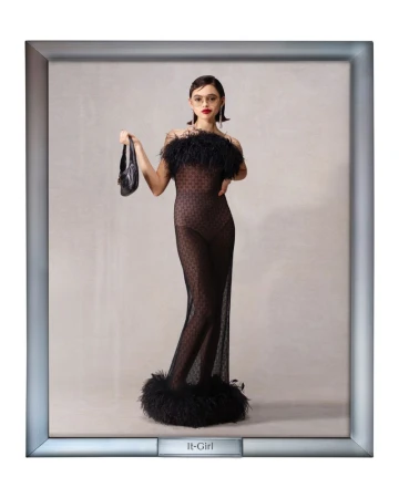Elegant woman poses in a black sheer dress with feather trim, holding a black purse, set against a neutral background inside a picture frame.