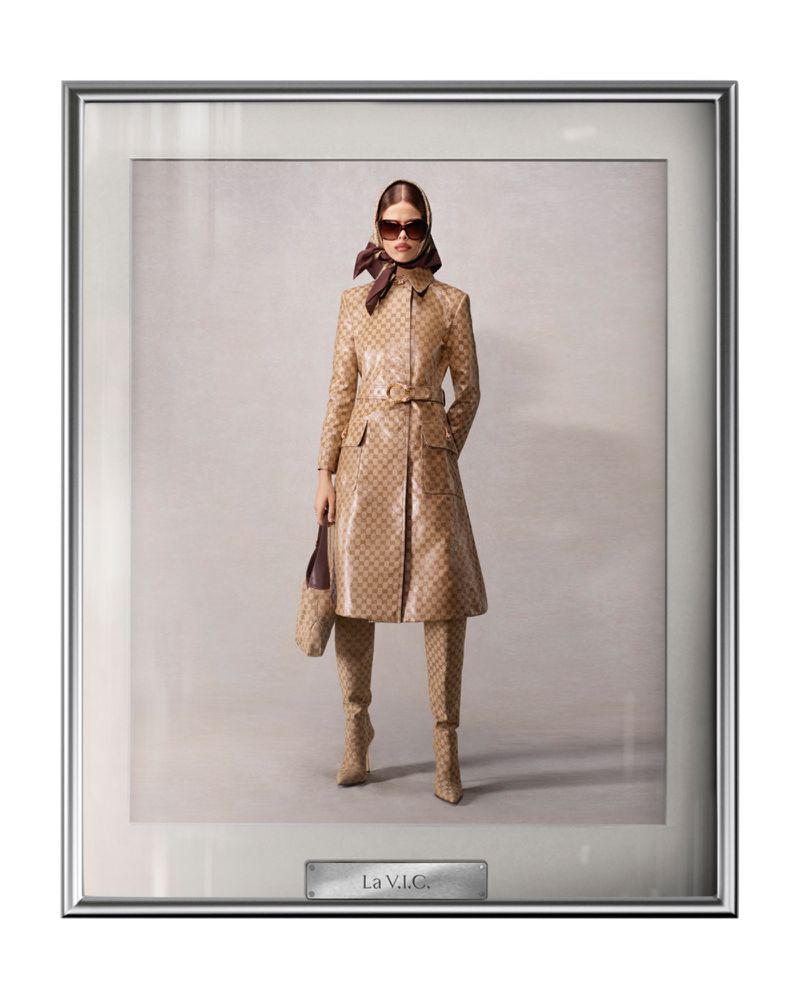 An adult female model poses in a framed photograph, wearing a tan patterned trench coat, matching pants and boots, a brown scarf and sunglasses.