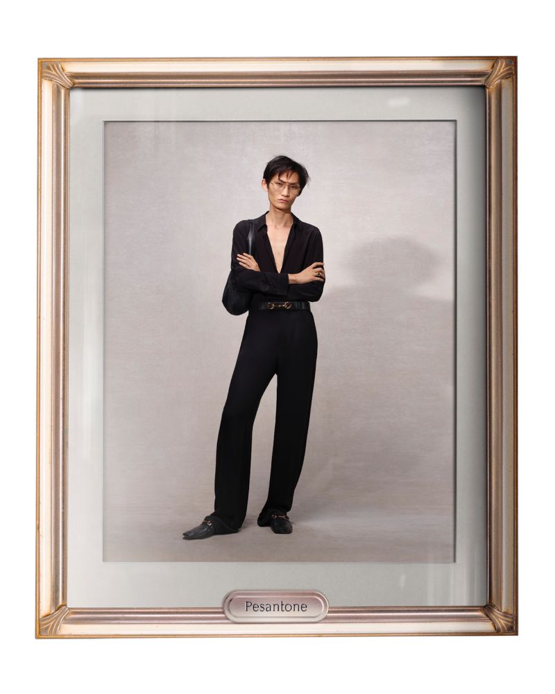 A person poses in a full-length shot within a decorative gold frame, wearing a black button-down shirt and black trousers against a light background.