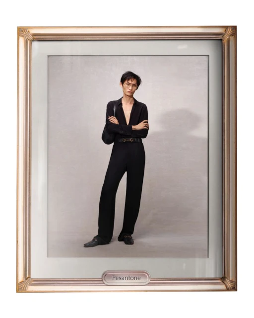 A person poses in a full-length shot within a decorative gold frame, wearing a black button-down shirt and black trousers against a light background.