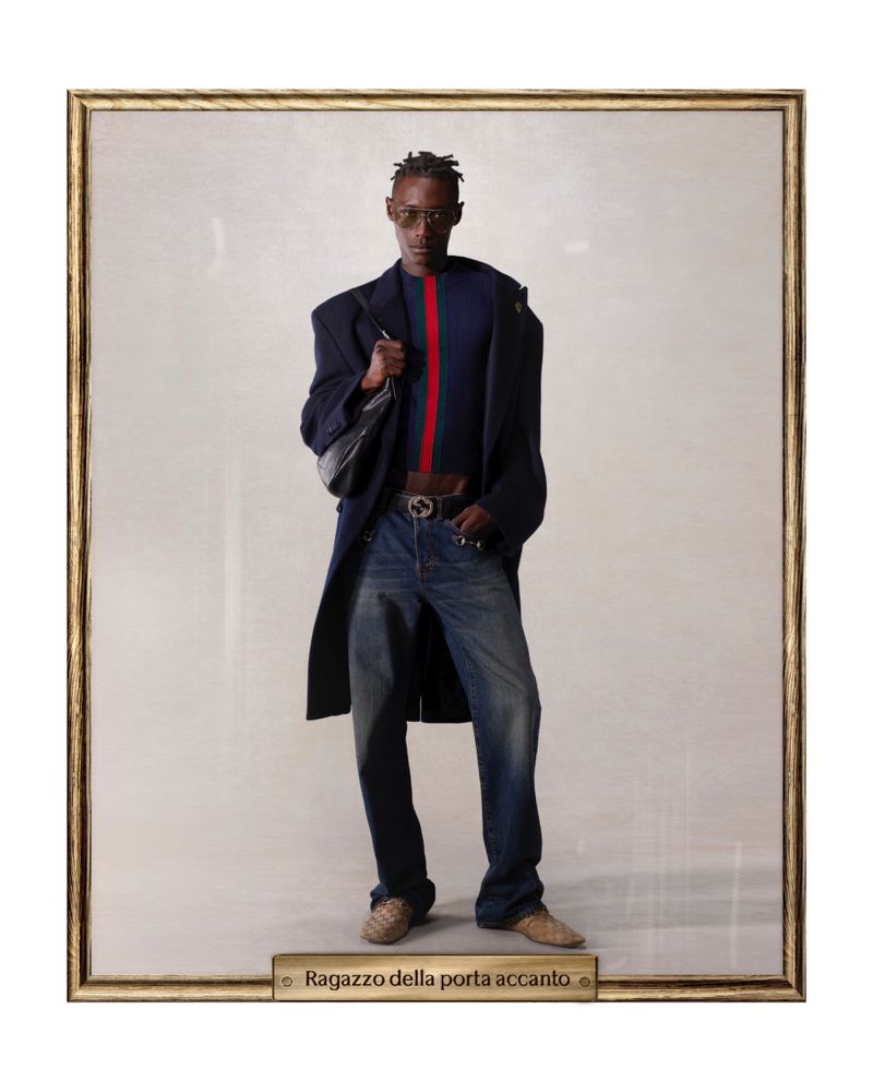 Framed studio portrait of a Black male model in a blue sweater and jeans, accessorized with a jacket, bag, and sunglasses.