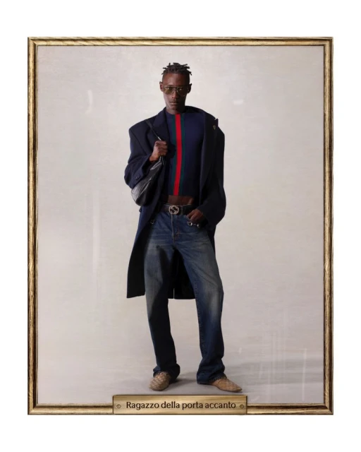 Framed studio portrait of a Black male model in a blue sweater and jeans, accessorized with a jacket, bag, and sunglasses.