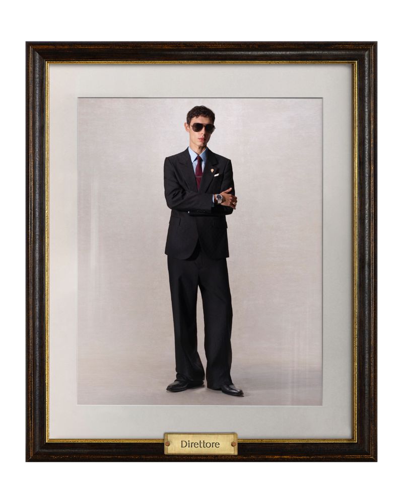 Framed full-length portrait of a young adult man indoors wearing sunglasses, dark suit, tie, and dress shoes against a neutral backdrop.