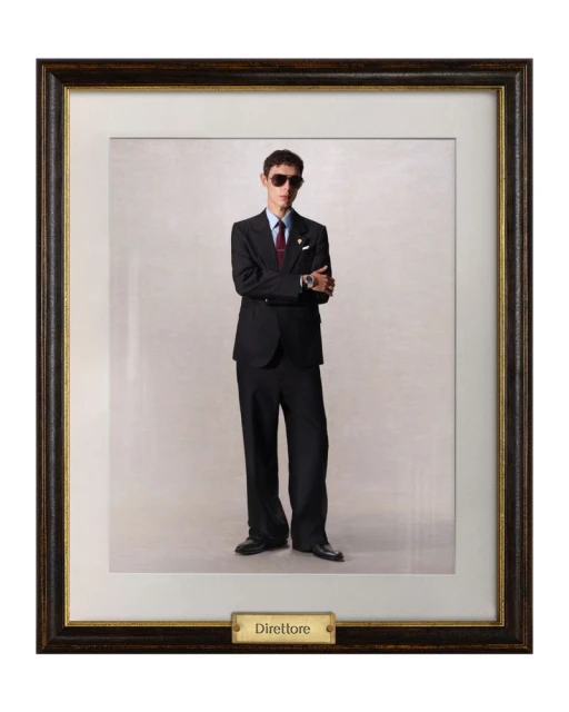 Framed full-length portrait of a young adult man indoors wearing sunglasses, dark suit, tie, and dress shoes against a neutral backdrop.