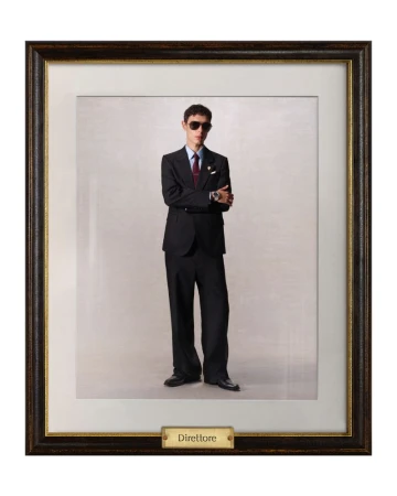 Framed full-length portrait of a young adult man indoors wearing sunglasses, dark suit, tie, and dress shoes against a neutral backdrop.