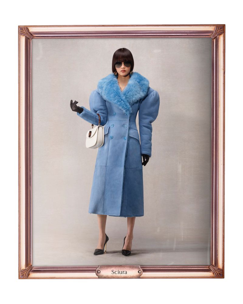 A stylish woman in a light blue shearling coat, black heels, and sunglasses, stands against a neutral backdrop in an ornate frame.