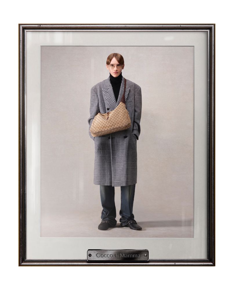 Framed fashion photograph of a slender adult male posing in a gray plaid coat with a woven tan shoulder bag, wearing a black turtleneck.