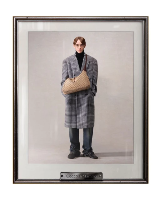 Framed fashion photograph of a slender adult male posing in a gray plaid coat with a woven tan shoulder bag, wearing a black turtleneck.