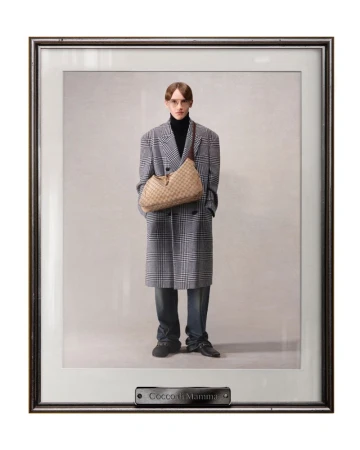 Framed fashion photograph of a slender adult male posing in a gray plaid coat with a woven tan shoulder bag, wearing a black turtleneck.