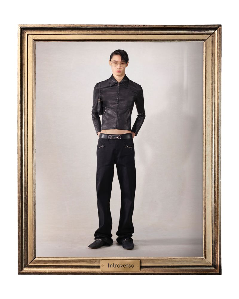 Full-length studio shot of an adult Asian male model in a black jacket and black trousers posing in a gold picture frame against a grey backdrop.