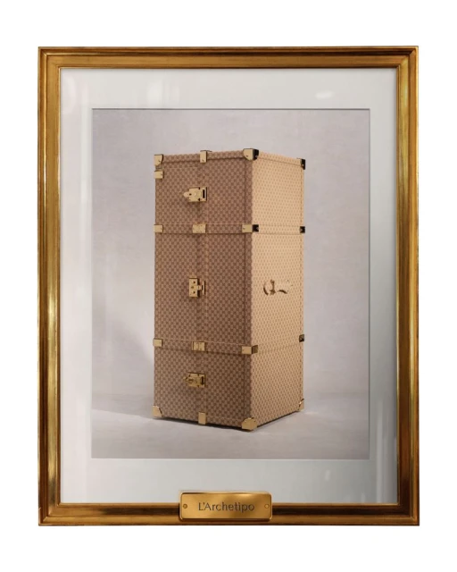 Framed photograph of a stacked set of tan and brown luggage with gold hardware and frame, indoors under studio lighting.