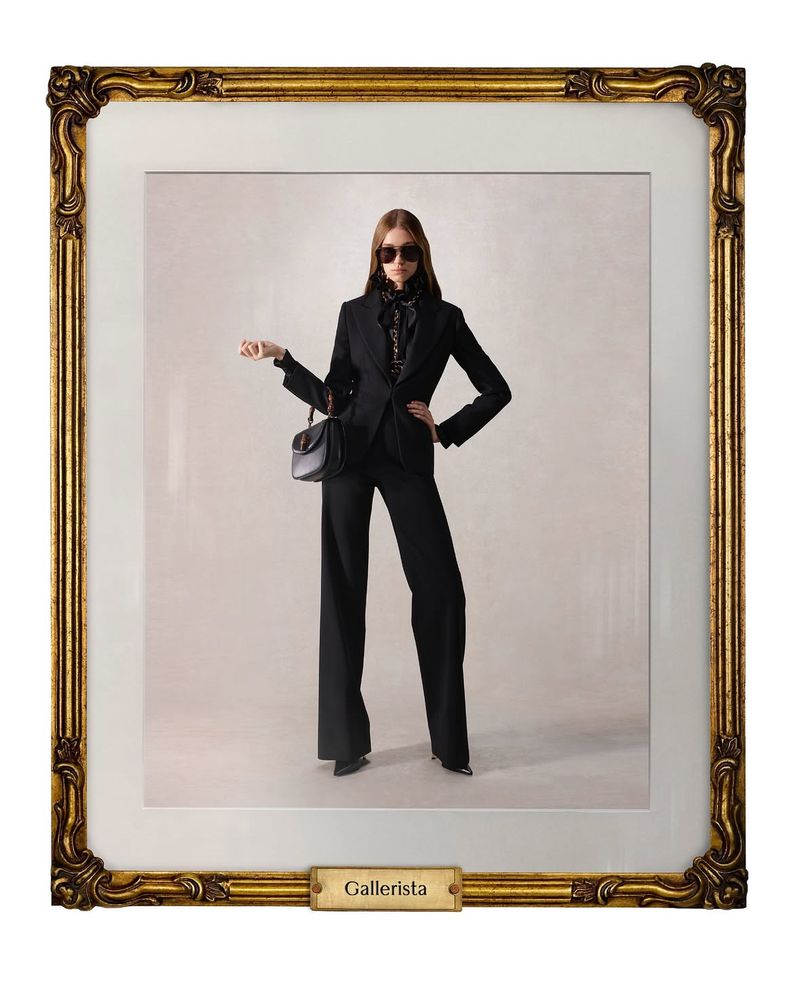 Framed studio shot of a stylish adult female wearing sunglasses and a black pantsuit, carrying a handbag, against a neutral background.