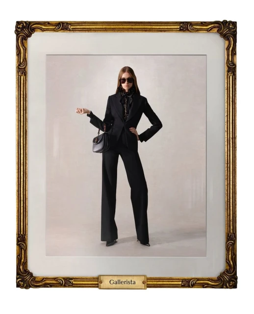 Framed studio shot of a stylish adult female wearing sunglasses and a black pantsuit, carrying a handbag, against a neutral background.