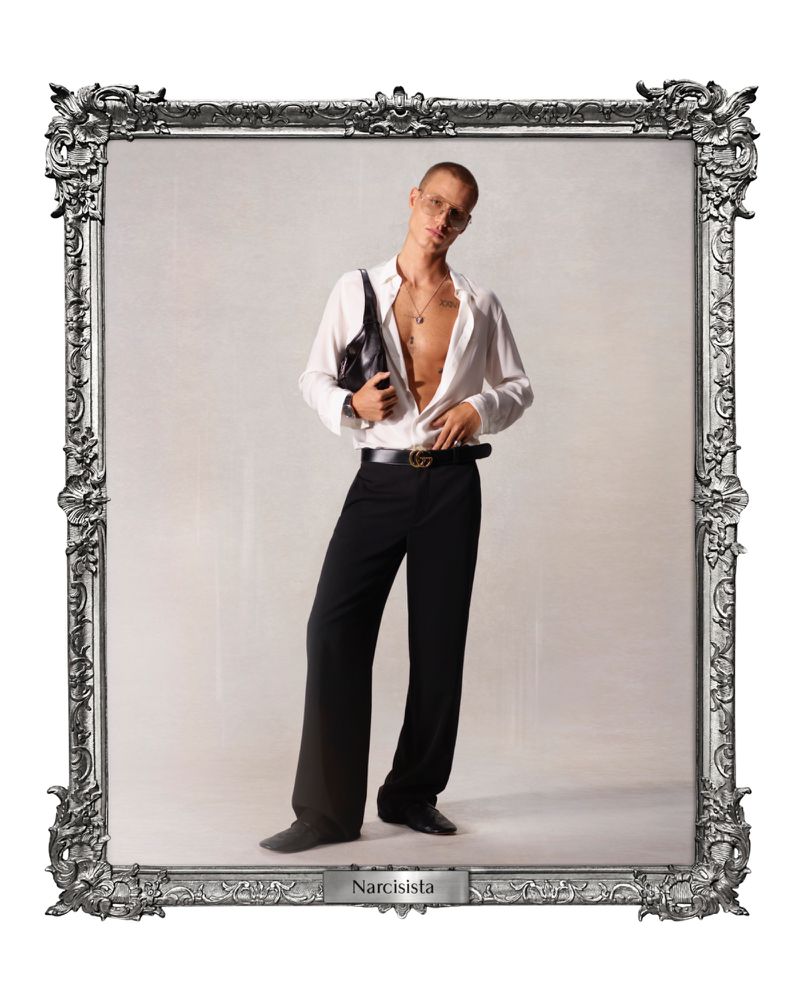 Full-length studio portrait of a young adult male in an unbuttoned white shirt and black trousers, accessorized with a handbag and jewelry.