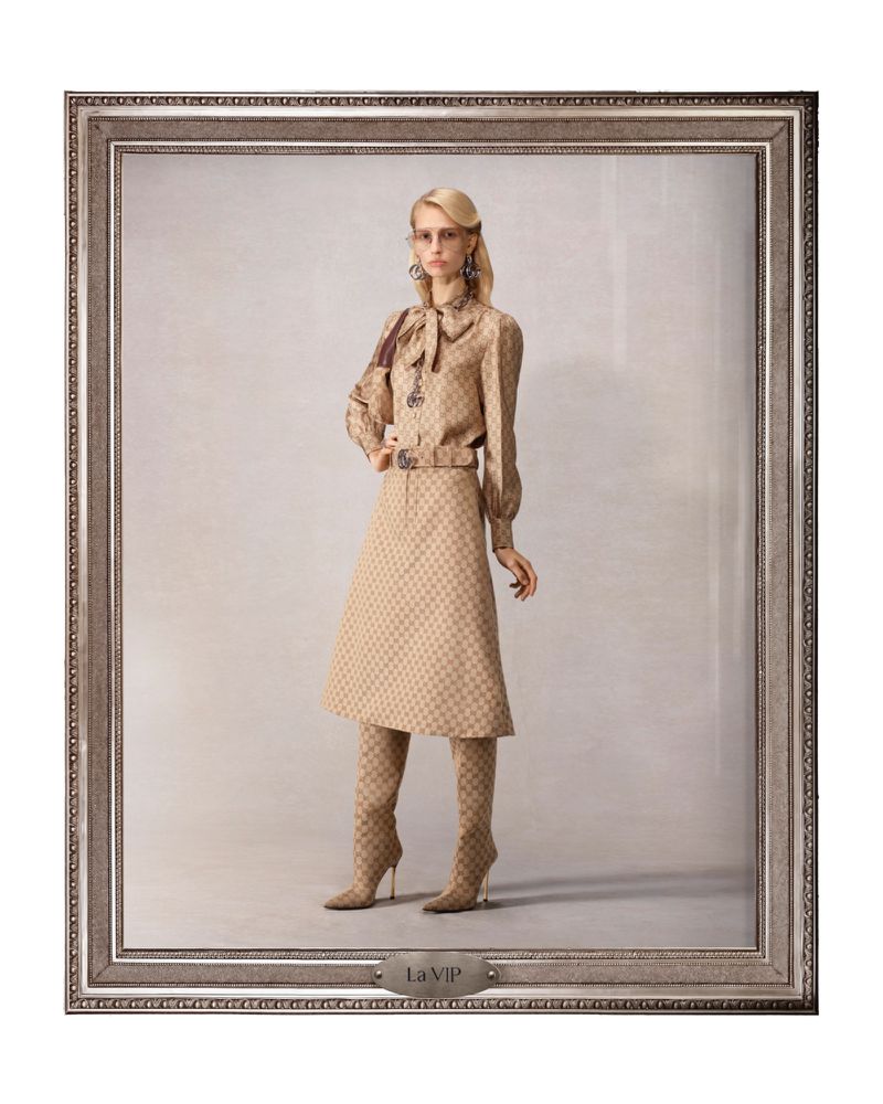 Full shot of an adult fashion model posing in a patterned tan long-sleeved shirt, skirt, and boots inside a decorative picture frame.