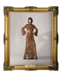 Model in ornate brown embellished dress standing inside a gilded antique frame against a neutral background