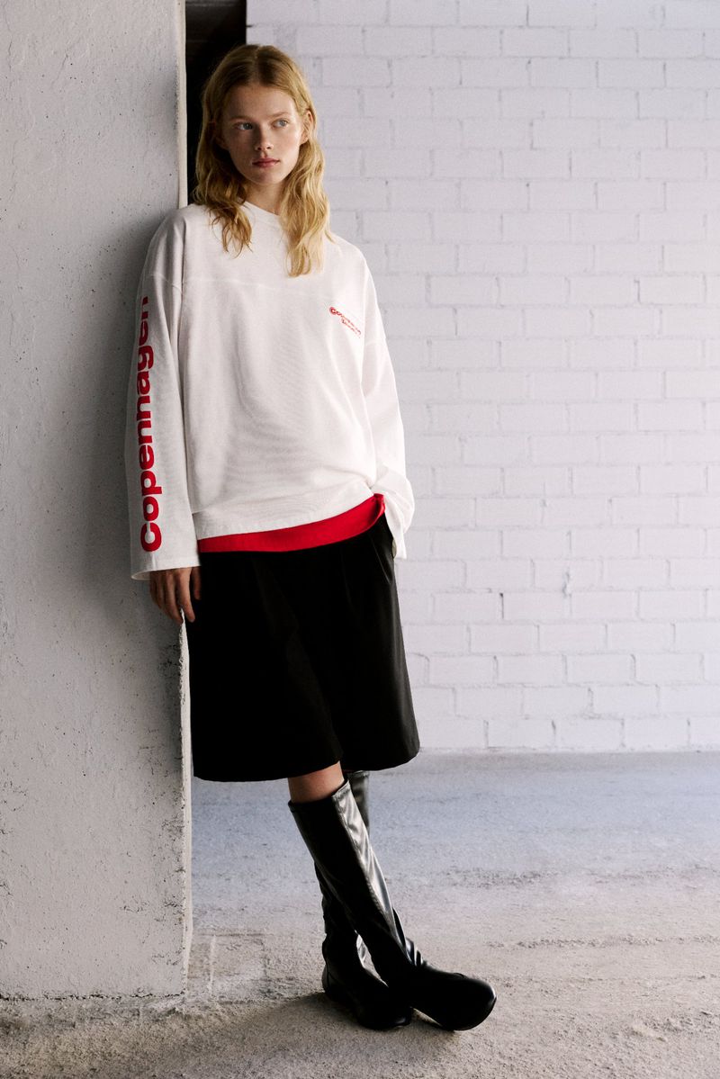Fashion shot of a young woman posing in a white long sleeve shirt, black skirt, and high boots against a white brick wall.