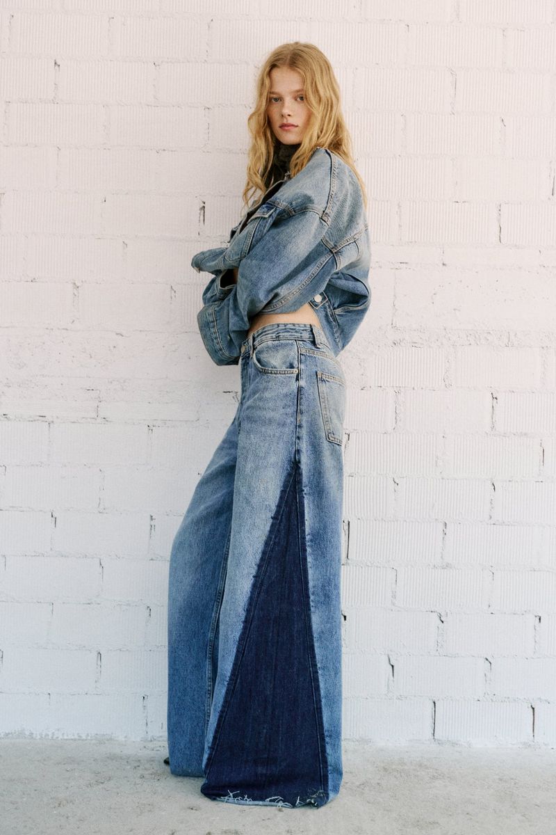A young woman with long, light blonde hair poses in a light blue denim jacket and wide-leg jeans against a white brick wall.