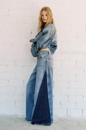A young woman with long, light blonde hair poses in a light blue denim jacket and wide-leg jeans against a white brick wall.