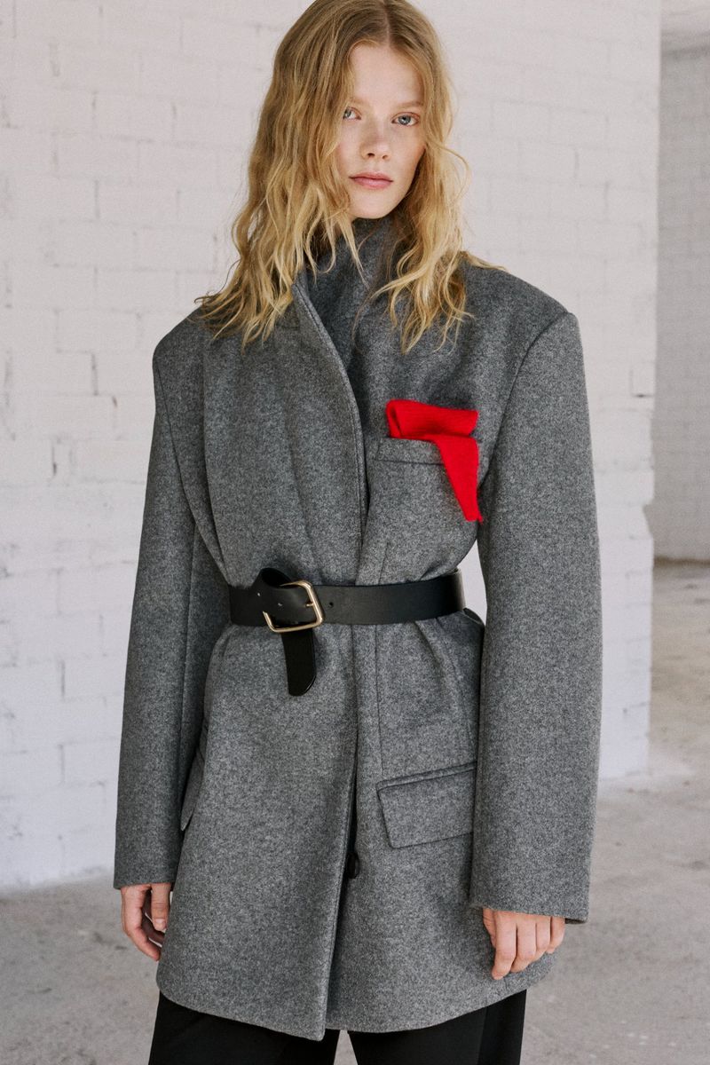 A fair-skinned woman with long wavy hair poses in a gray wool coat cinched with a black belt, standing against a white brick wall.