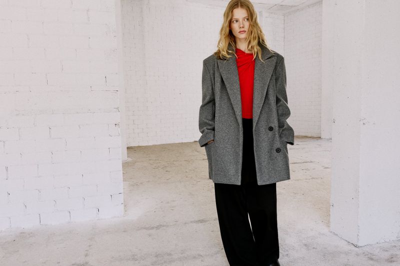 A fair-skinned woman with long blonde hair stands indoors wearing a gray overcoat and black pants, posing in a minimalist style.