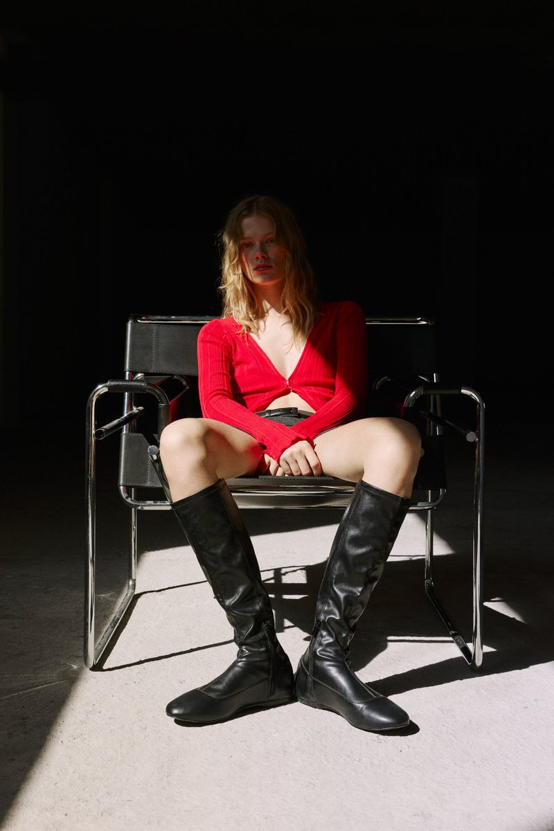 A blonde woman sits in a black chair, wearing a red cardigan, black shorts, and black boots, in a studio setting with a dark background.