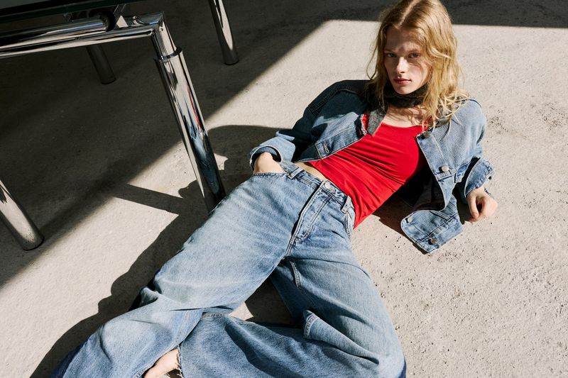 Model Sophia Enggaard wearing wide-leg jeans, a denim jacket, and a bright red top, reclining on a concrete floor in hard sunlight.