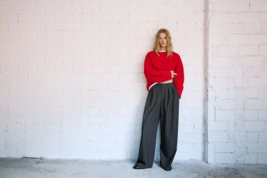Model standing against a white painted brick wall wearing a bright red sweater and dark wide-leg trousers.