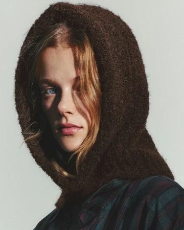 Close-up shot of a blonde woman wearing a brown knit balaclava and a green and red plaid shirt against a neutral background.