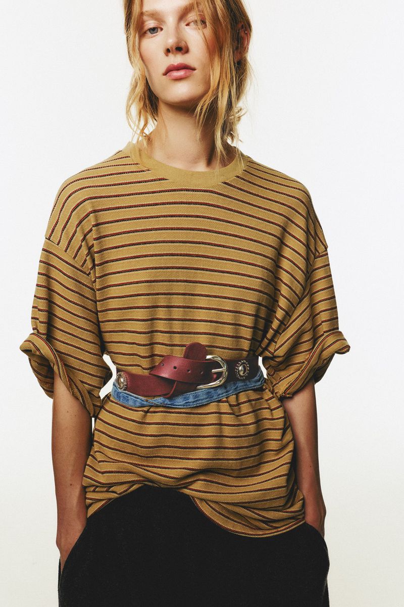 Adult woman poses in a tan and burgundy striped t-shirt with a brown leather and blue denim belt, against a white backdrop.