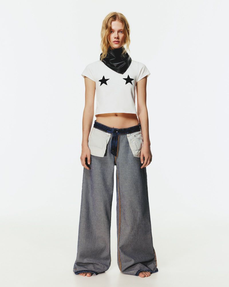 Full shot of a blonde woman wearing a white crop top with stars, wide-leg jeans, and a black scarf, posing in a brightly lit studio.