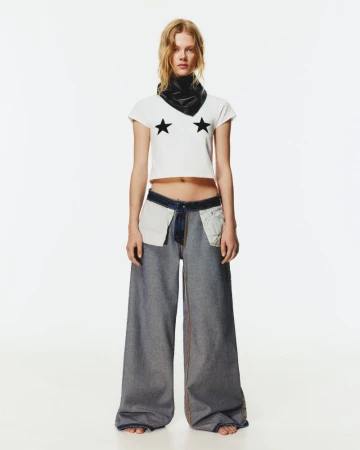 Full shot of a blonde woman wearing a white crop top with stars, wide-leg jeans, and a black scarf, posing in a brightly lit studio.