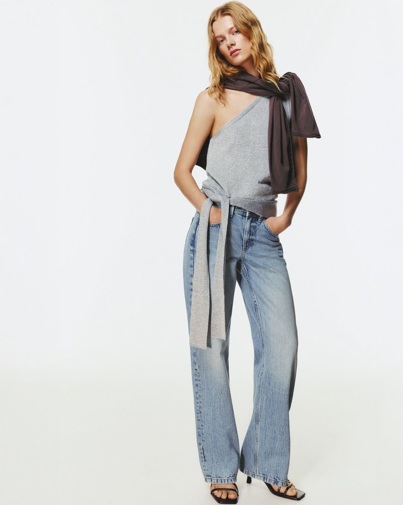 Fashion model poses in a studio wearing a gray top, blue jeans and black heels. The image is shot in a bright photo studio.