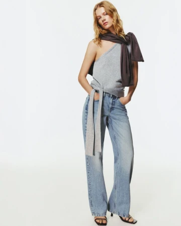 Fashion model poses in a studio wearing a gray top, blue jeans and black heels. The image is shot in a bright photo studio.