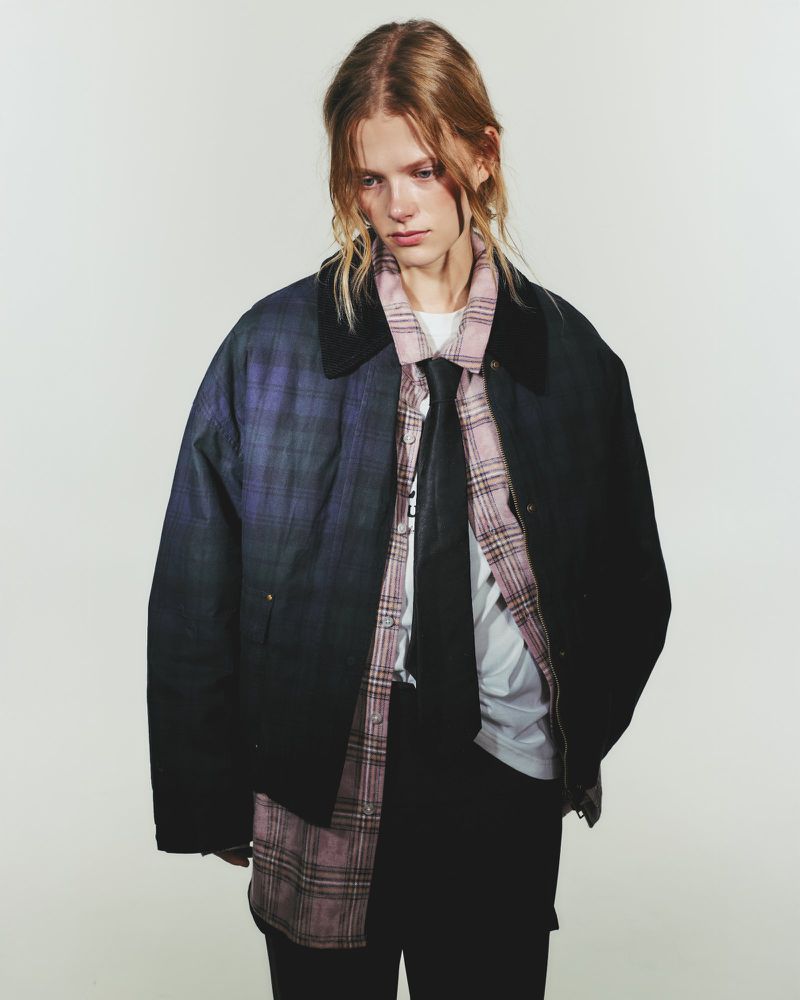 A person in a dark jacket poses indoors in a studio with a plaid shirt and black pants in a medium shot with neutral lighting.