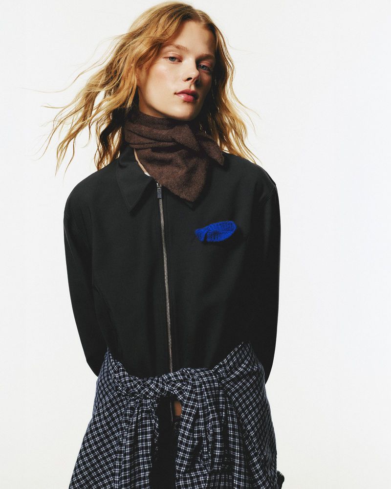 Fashion portrait of a blonde female adult wearing a black jacket with scarf, flannel tied around waist against a white backdrop.