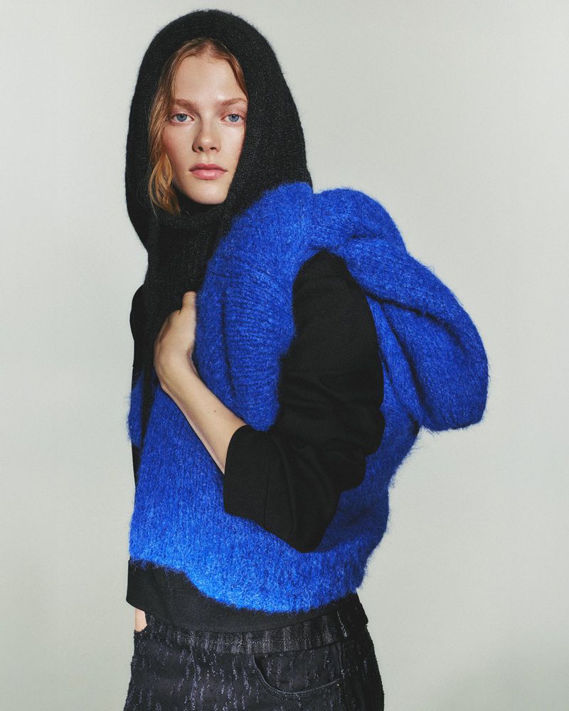 A blonde woman poses indoors wearing a black hoodie and a blue, fluffy vest. She looks at the camera with a neutral expression.