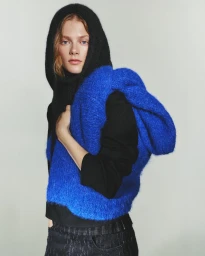 Model wearing a black hooded sweater and vibrant blue knit sweater draped over her shoulders against a pale background