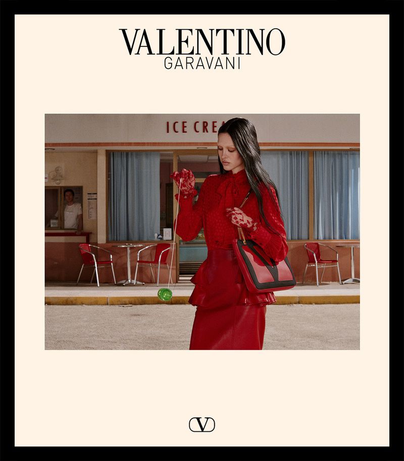 A model in a red outfit stands in front of an ice cream shop with a red Valentino Garavani purse, promoting high fashion.