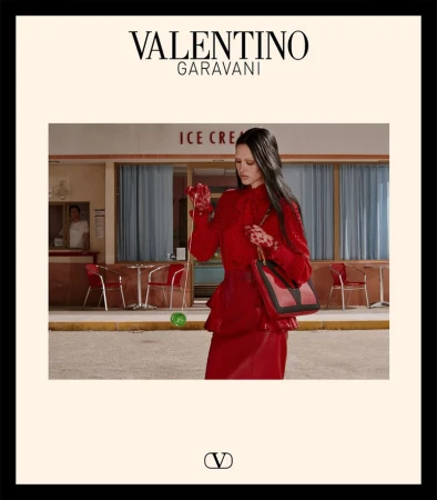 A model in a red outfit stands in front of an ice cream shop with a red Valentino Garavani purse, promoting high fashion.
