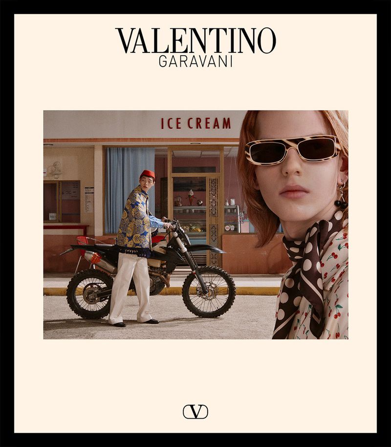 Fashion editorial shot: woman in polka dot scarf, sunglasses. Man on motorcycle near ice cream shop, wearing patterned jacket.