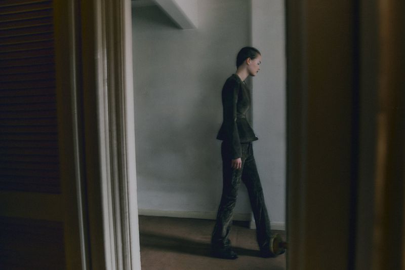 A slender woman in a dark peplum top and pants walks in a hallway, viewed through a doorway; muted colors, natural light.