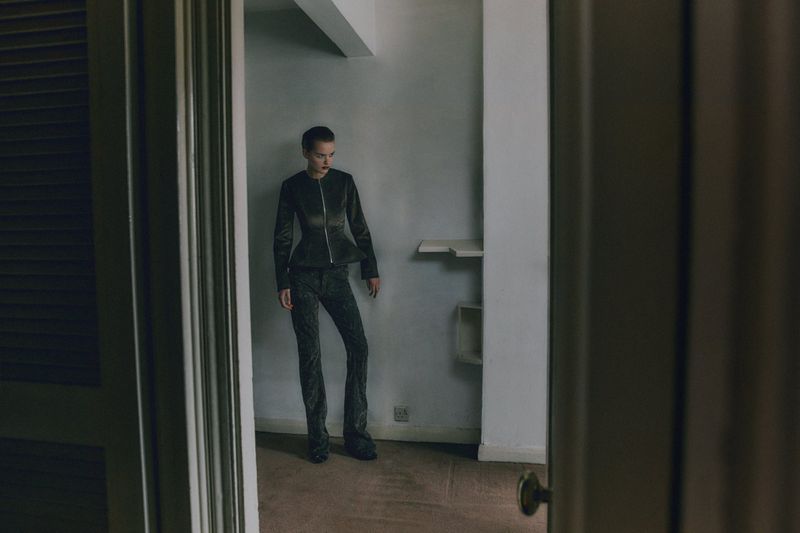 An adult female wearing a dark jacket and trousers stands in an empty room, seen through a doorway, in a fashion-editorial composition.