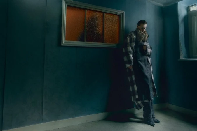A person in a coat stands in a dim room next to a window, creating a contemplative or somber mood with a sense of isolation.