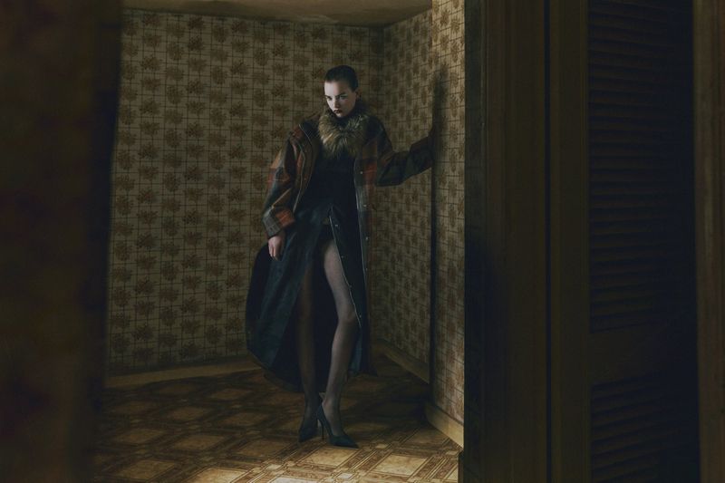 A tall, slender woman in a long coat poses in a corner of a room with patterned wallpaper and flooring, casting a mysterious, elegant shadow.