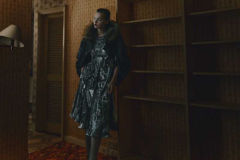 Fashion editorial shot of a stylish woman in a sequined dress and jacket with a fur collar, posing in a dimly lit interior.
