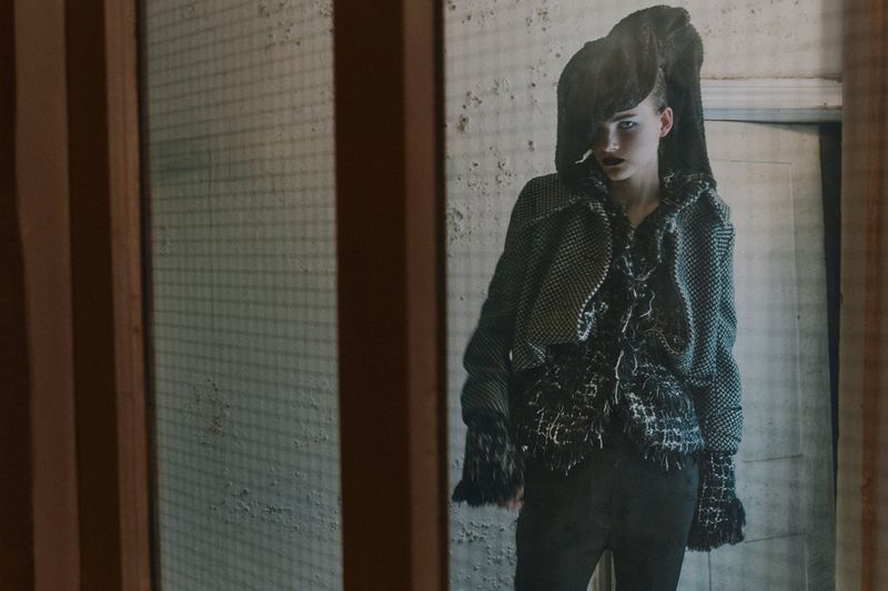 Indoor fashion shot of a model in a gray jacket, dark top, and unique headwear, posing in front of a textured wall and weathered door.