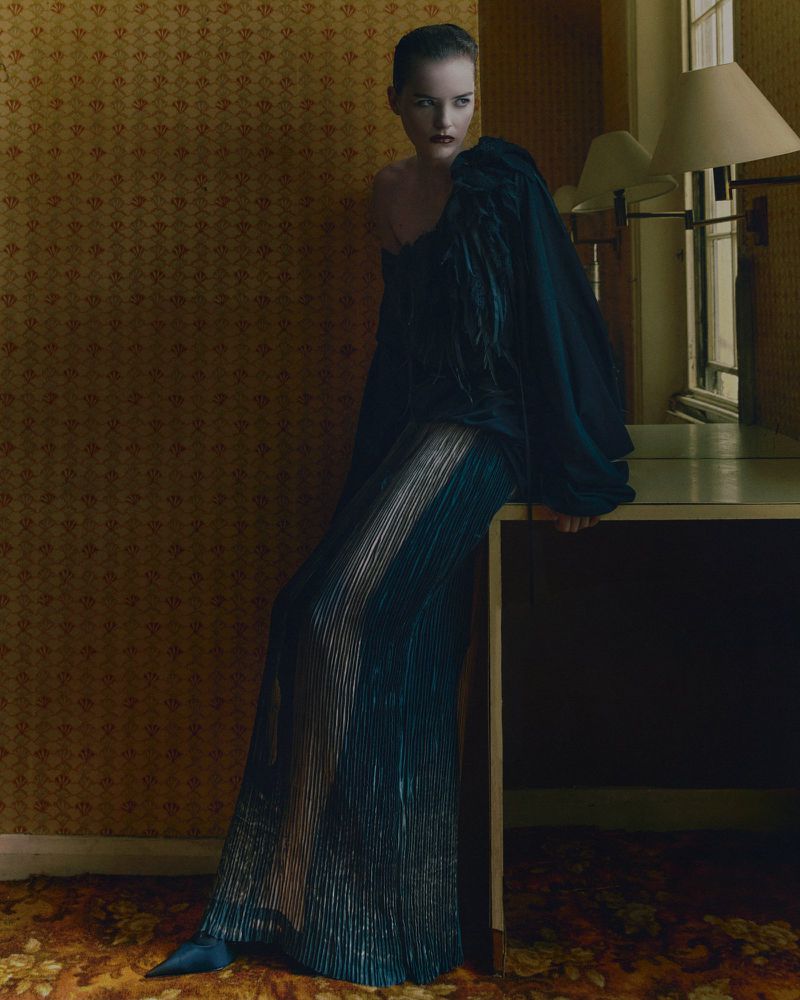 Elegant woman in dramatic black gown with feather detail, seated on a table, set against a warm-toned, patterned wall in a stylish indoor setting.