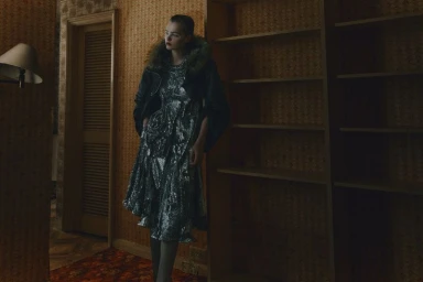 Model standing in a dimly lit room wearing a metallic patterned dress and dark jacket with a fur collar